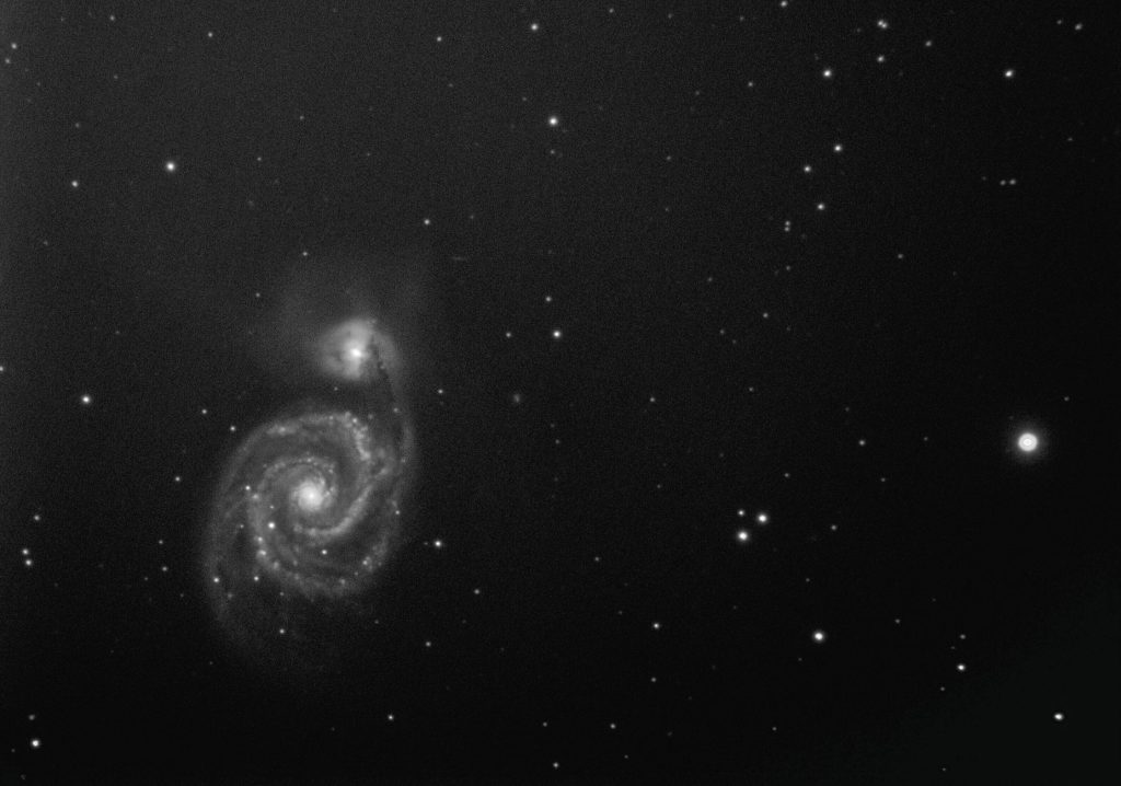 Imagem em preto e branco de uma galáxia espiral vista no espaço profundo, cercada por um campo de estrelas.